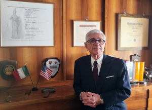 Alexander Guttieres, Of Counsel at Perkins Law Offices, standing in an office with legal certificates, flags of Italy and the USA, reflecting his international legal expertise.